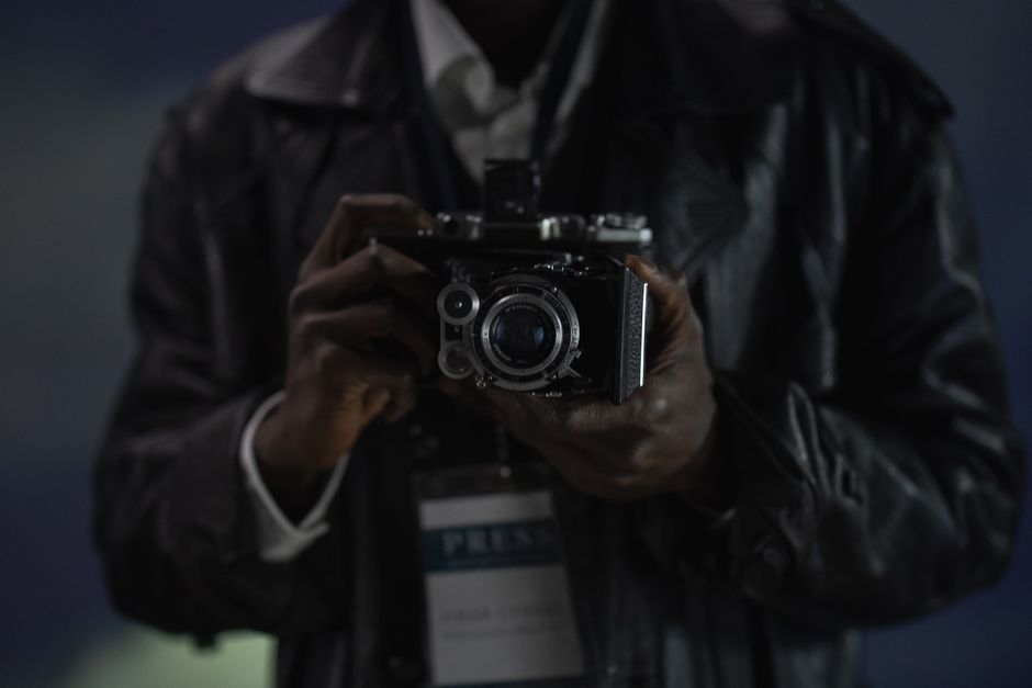 A close-up photograph of a person holding a vintage camera, possibly a reporter.